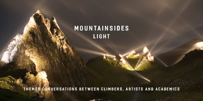 A mountainscape at night artificially lit by bright spotlights. White light beams project up into the night sky. 